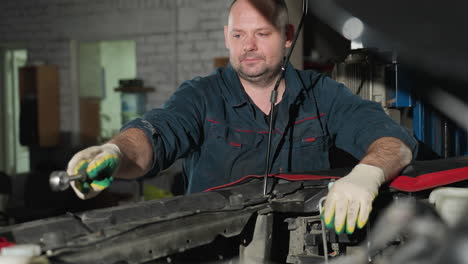 mechanic in blue uniform with gloves working on car engine, exchanging tools with colleague in background, workshop setting with various tools and equipment visible