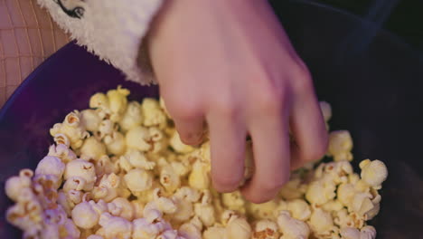 close up of female hand reaching into bowl of popcorn, fingers selecting pieces while seated beside visible leg in fishnet pantyhose under soft ambient lighting in cozy setting