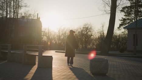 backshot of a woman wearing a brown coat, jeans, and white shoes, carrying a backpack as she walks alone through a park. the warm, golden light casts long shadows and creates a serene atmosphere