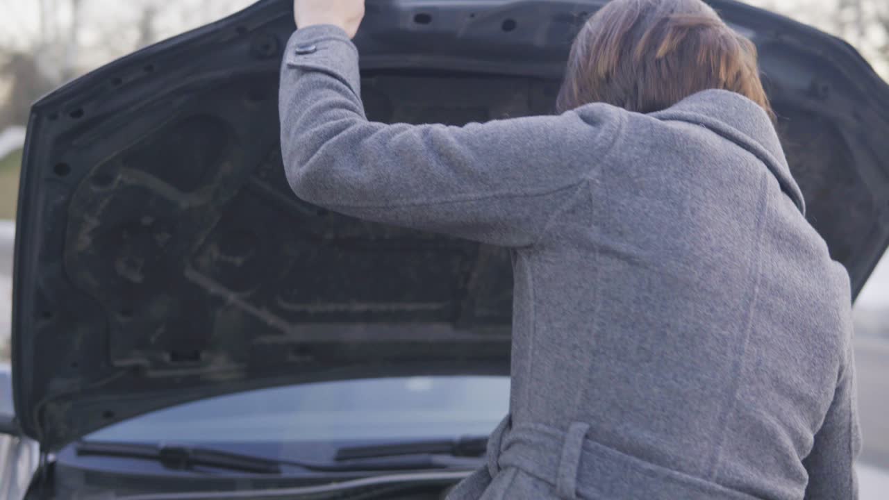Attractive business woman in grey coat opening the car's bonnet and looking under the hood in the street
