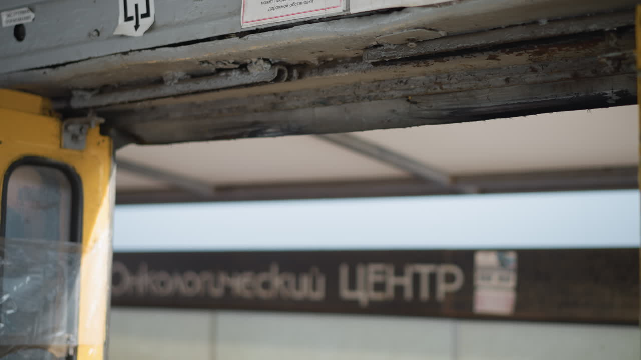 close view of vehicle door mechanism opening and closing at stop, worn metal track and yellow frame move in winter light, blurred city shelter signage outside, subtle vibration