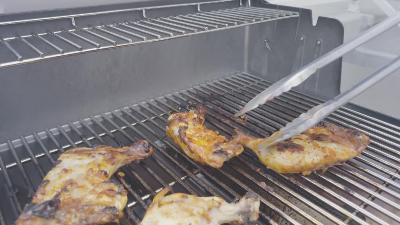 Grilled chicken legs cooking on a stainless steel BBQ grill. Stainless steel tongs turn the chicken as the camera pulls back on an angle across the grill