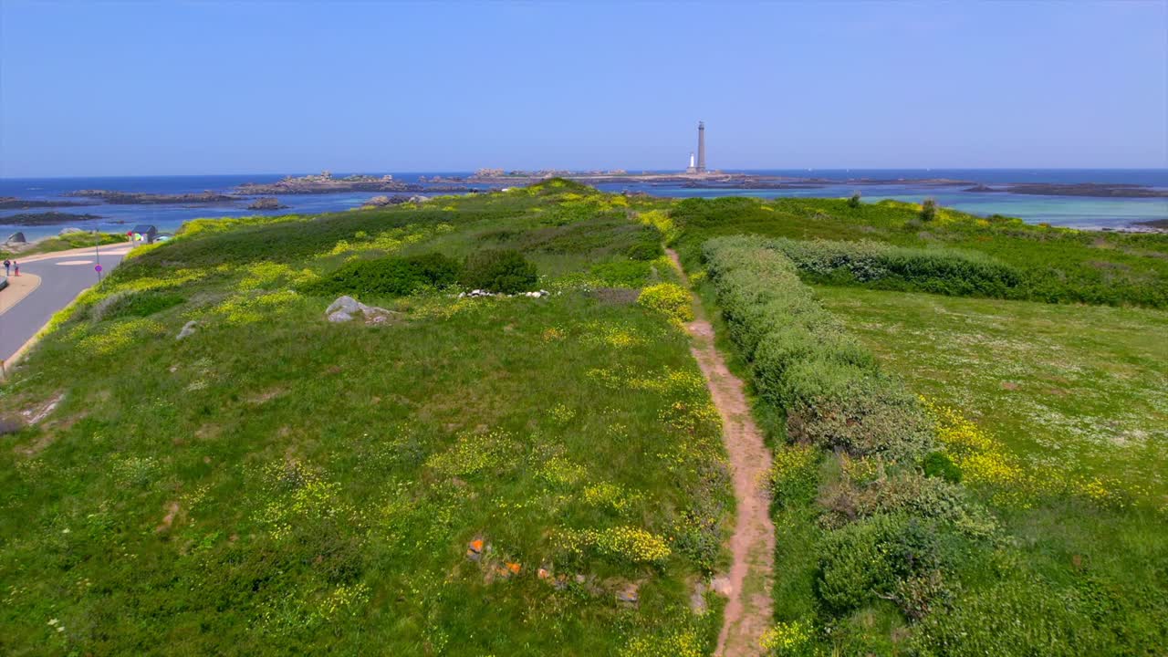 A scenic coastal path lined with green vegetation and yellow wildflowers, with the iconic lighthouse in the distance near the coastline in Brittany, France. Aerial view captures the natural landscape.