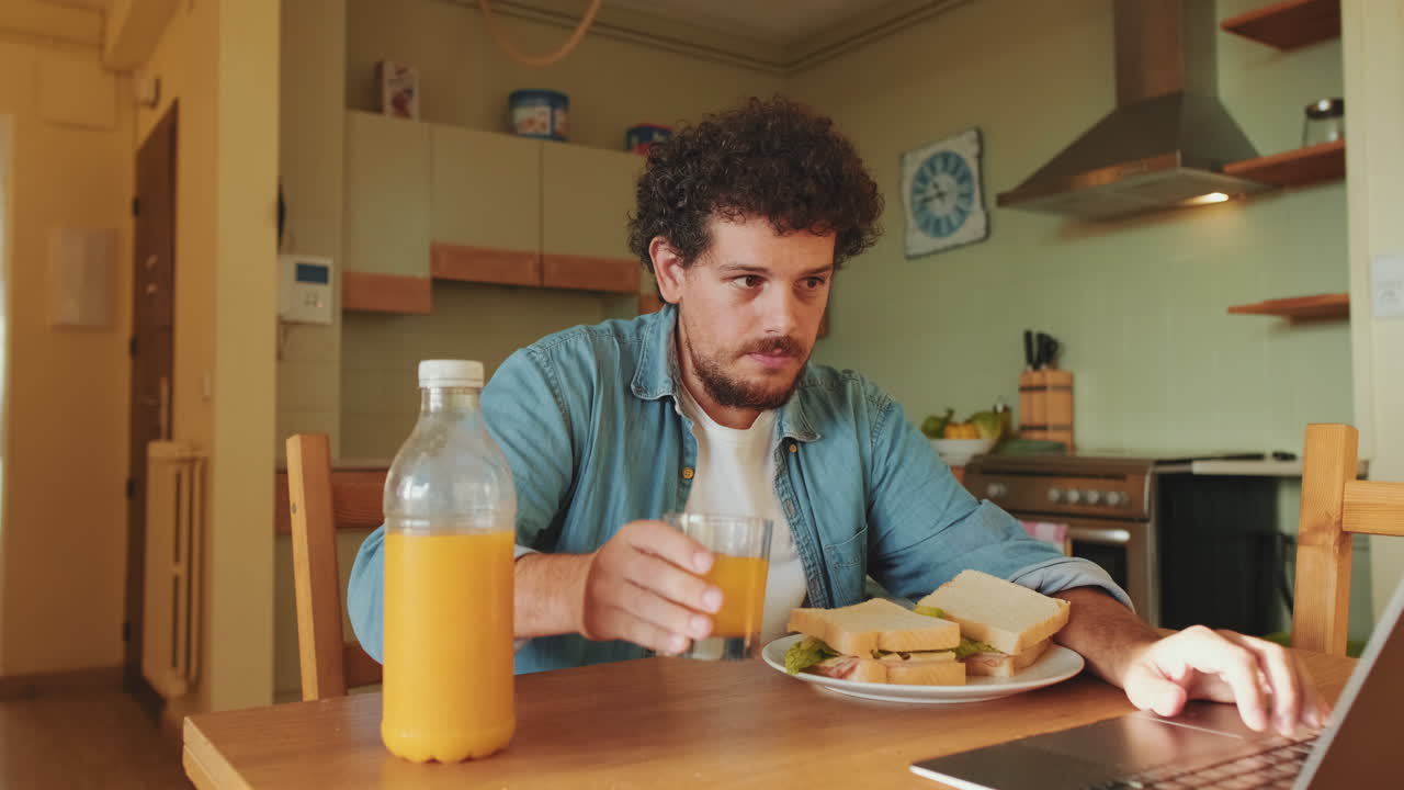 Man eating sandwich and drinking orange juice while working on laptop in kitchen