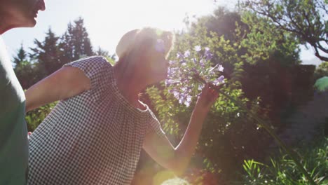 Happy-caucasian-senior-couple-walking-in-garden-in-the-sun-the-woman-smelling-a-flower