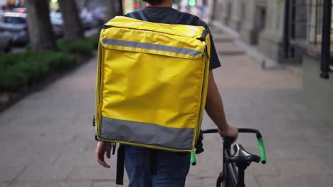 Delivery-man-walking-alone-with-bike-and-yellow-bag,-rear-view