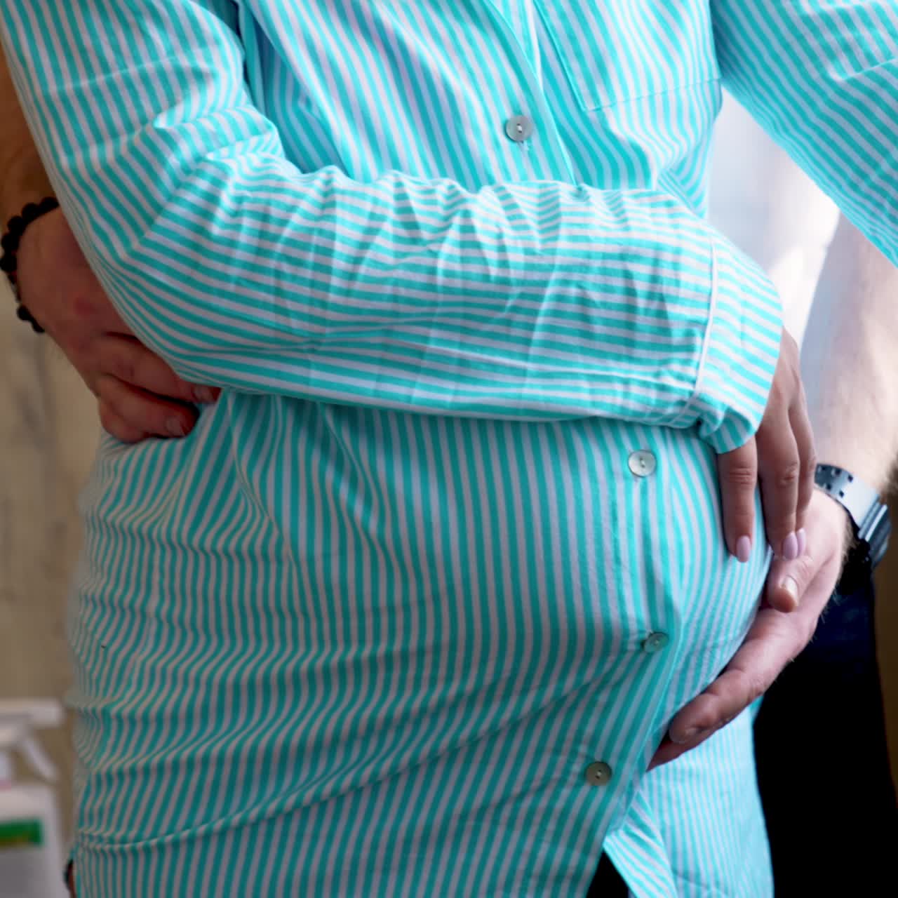 Unrecognized pregnant woman stands at the window-sill holing by the belly. Husband strokes her low back and abdomen standing behind