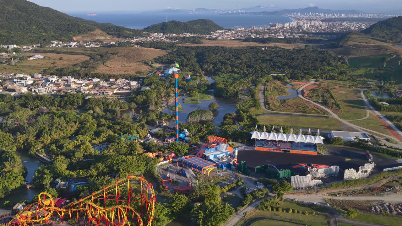 Drone view above Beto Carrero World in Penha, Brazil, showing colorful roller coasters, tall drop tower, water canals, and themed architecture amid green surroundings and distant urban skyline