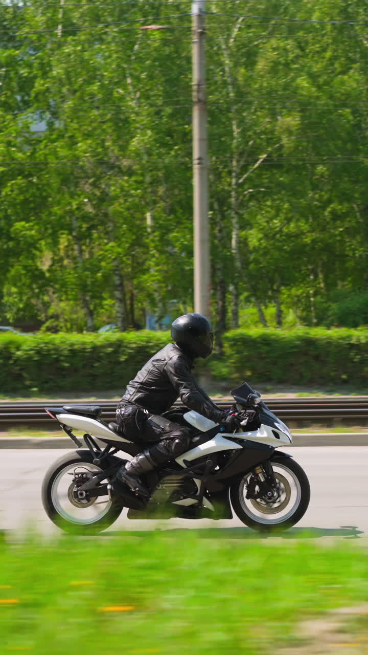 Skilled rider in black leather uniform and helmet drives powerful bike along large road past green park in city on sunny day side view slow motion