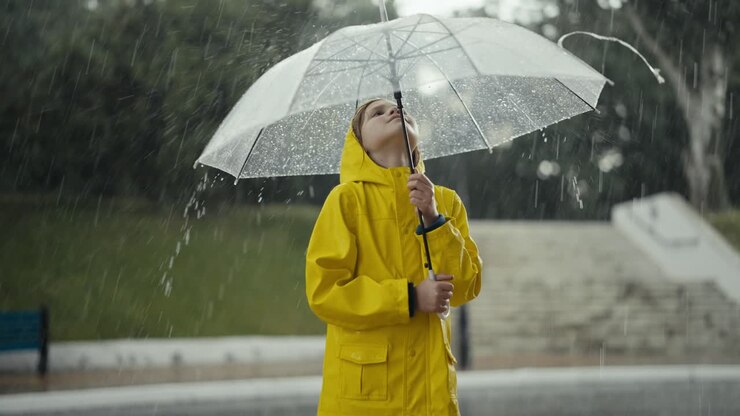 Happy teenage girl in a yellow jacket holds an umbrella and watches water drops flow down it during heavy rain while walking in the park