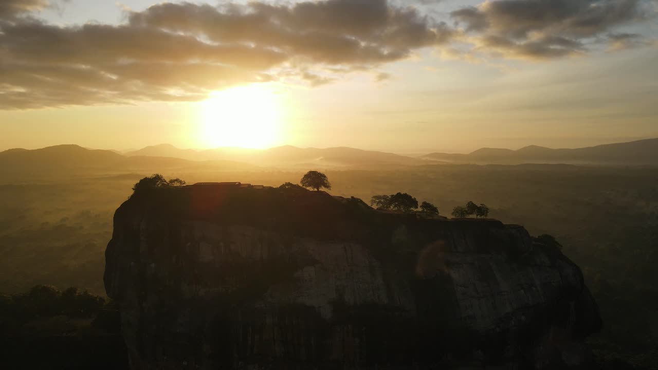 Beautiful yellow sunset just above the silhouettes of the mountains with tall trees in the foreground on top of the citadel called Sigiriya in Sri Lanka on a partly cloudy day. Wide drone panning shot