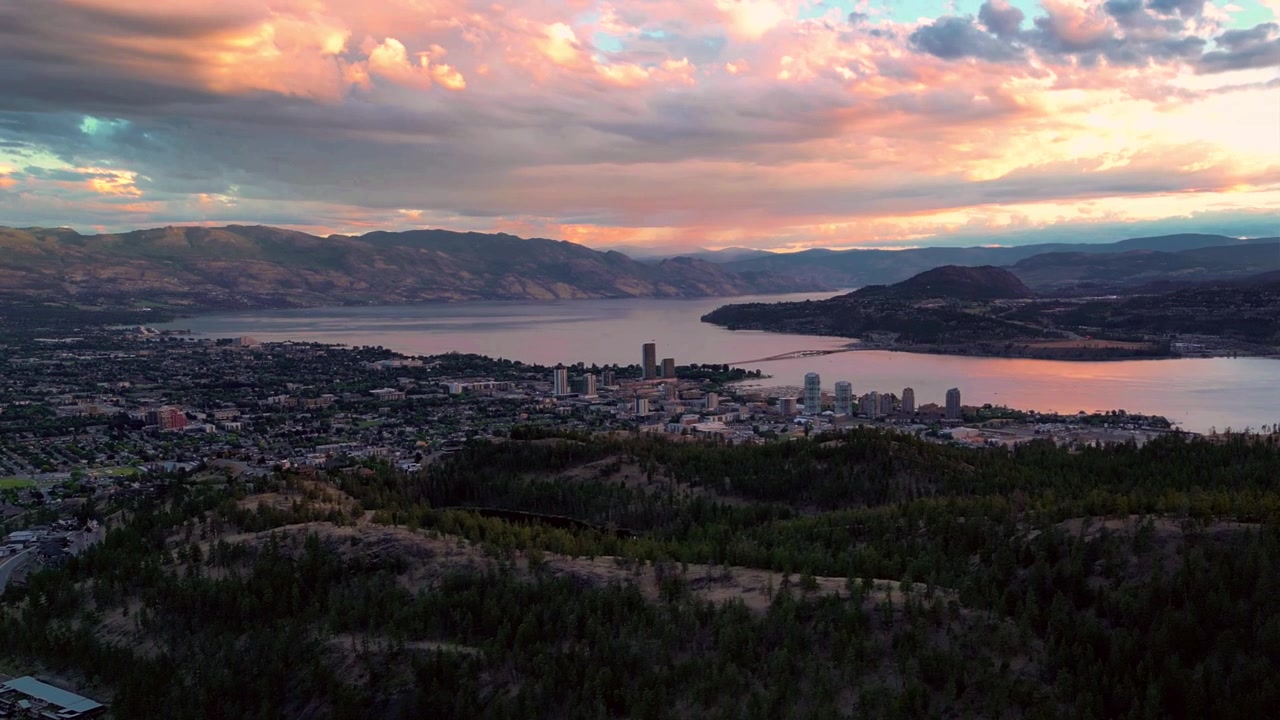 Ripresa aerea cinematografica del panorama della città di Kelowna al tramonto, che mostra il Monte Knox in primo piano, il lago Okanagan e il ponte sotto un cielo crepuscolare rosso e drammatico.