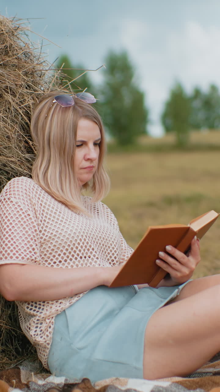 Young woman sitting on blanket, resting against hay bale, flipping book pages thoughtfully, green notebook beside her on grass, blurred rural background with trees and hay bales in distance