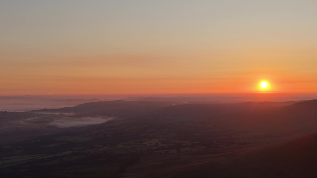 Vista aérea sobre los campos de Brecon Beacons en el sur de Gales, con el cálido resplandor del amanecer llenando un valle neblinoso.