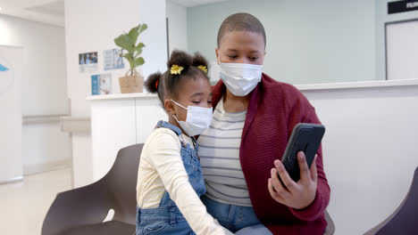 African-american-mother-and-daughter-wearing-face-masks-using-smartphone-in-hospital,-slow-motion