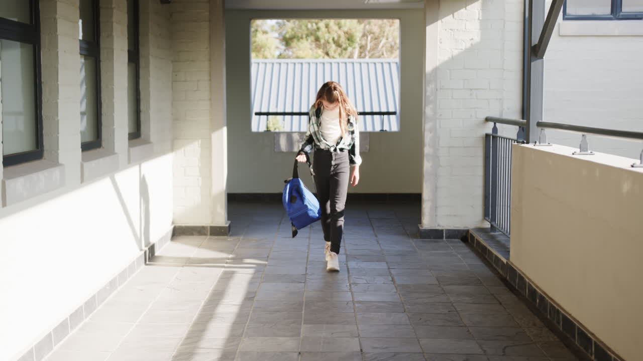 Young girl walking in school hallway carrying blue backpack, looking down