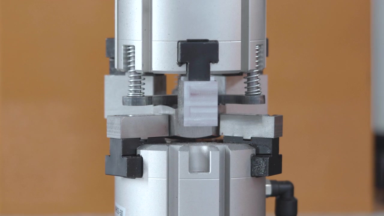 Silberner industrieller Klemmmechanismus, zentriert vor warmbraunem Hintergrund, mit bearbeiteten Metallblöcken, schwarzen Seitengriffen, sichtbaren Federn und zylindrischem Sockel in klarer Vorderansicht unter weichem Licht