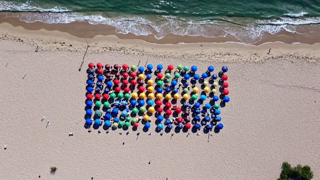 Tourists relaxing under colorful beach umbrellas arranged in a geometric pattern on a sandy beach, creating a vibrant and cheerful scene from an aerial perspective