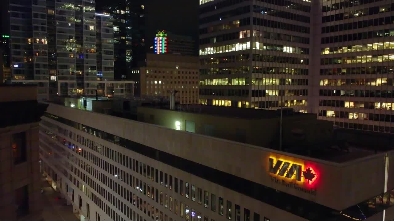 Drone flies above stunning glass buildings with the Via logo at night, with city lights in Montreal, Canada.