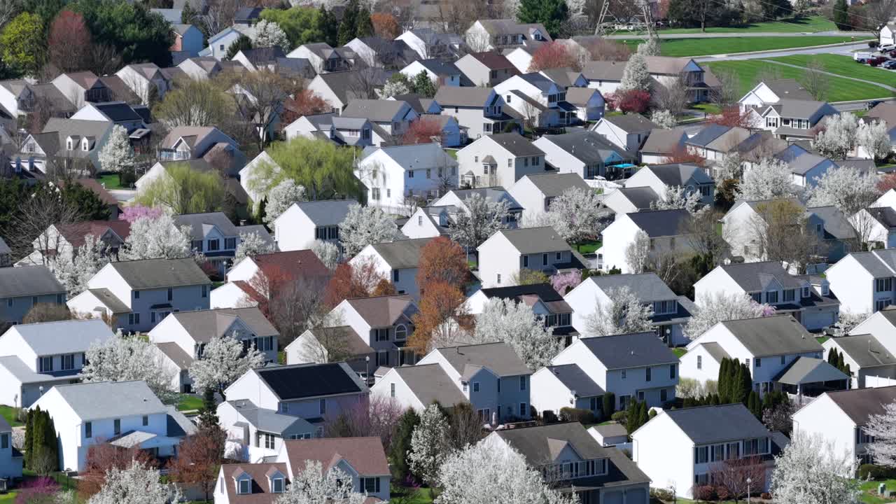 春のアメリカ郊外住宅地の空撮。住宅が立ち並び、木々が咲き誇り、柔らかな日差しが差し込む。穏やかで活気あるコミュニティの雰囲気。広角ショット。