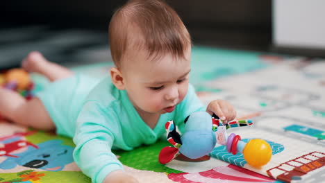 lovely baby boy in blue romper lies on colorful mat. cute kid playing with a toy, pulling it to his mouth and looking at it with interest.