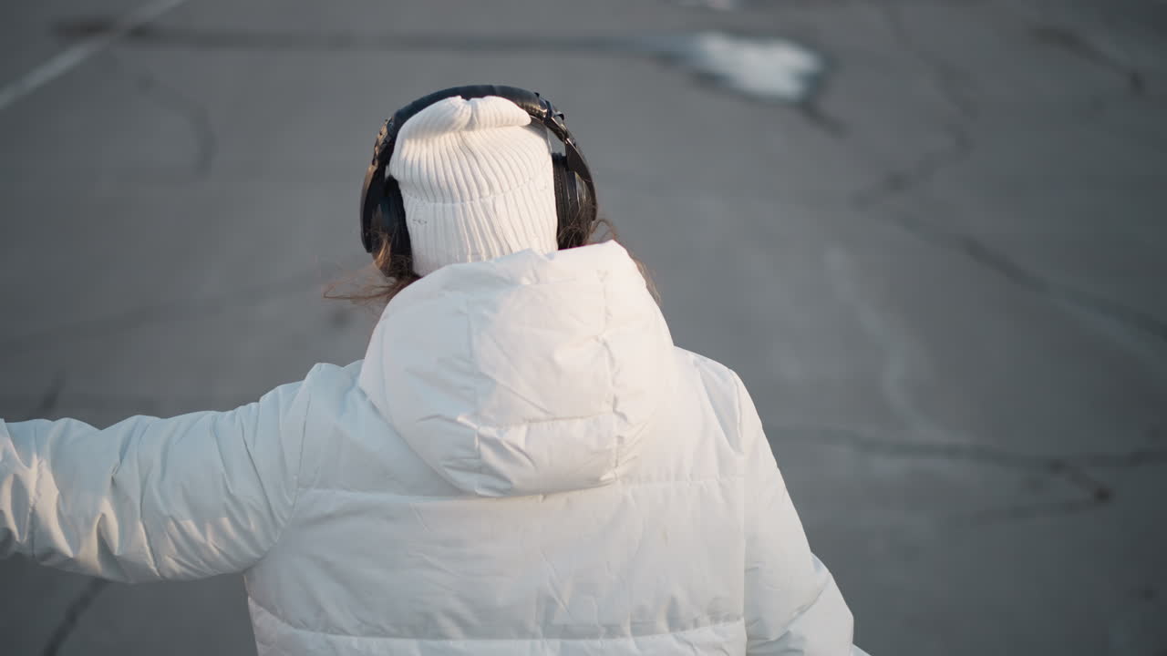 Back view of creative dancer twirling gracefully while moving along urban tiled walkway on cracked pavement, dressed in white winter jacket, headphones, and beanie, enjoying rhythm on cold sunny day