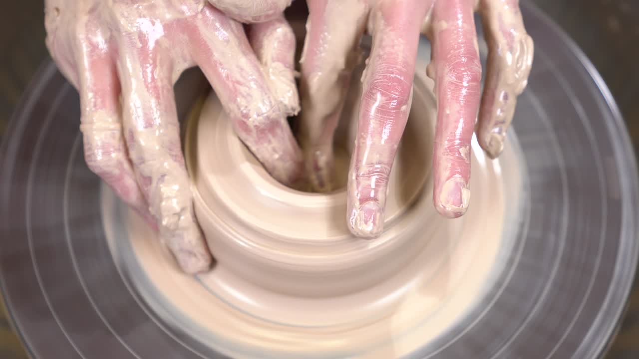 Muddy potter hands are shaping a clayware from the lump of clay on the spinning potter's wheel.