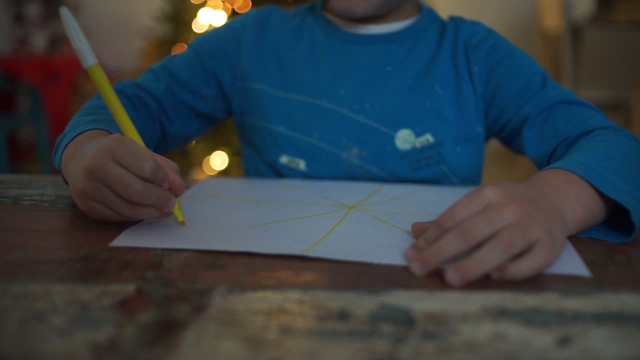 Footage of a kid drawing at paper with a christmas tree on the background