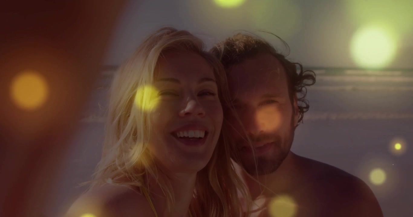 Coppia che crea contenuti di marketing di viaggio su una spiaggia sabbiosa, con sovrapposizione di sfere bokeh illuminate dal sole.
