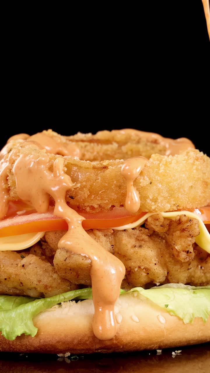 Primo piano di salsa Thousand Island versata su un hamburger di pollo fritto con anello di cipolla, lattuga, pomodoro e formaggio, sotto illuminazione da studio