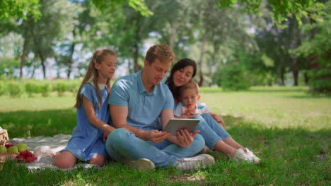 young family sitting with tablet on sunny park. happy people use laptop outdoors