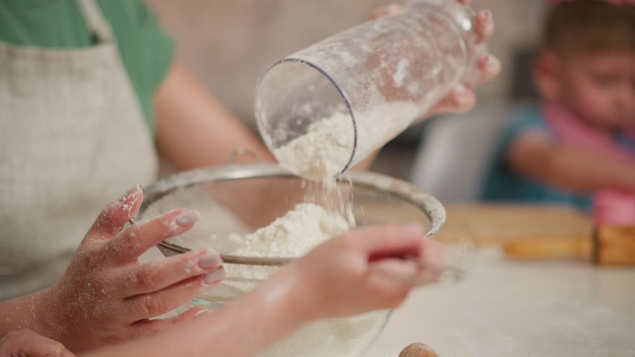 close up of child flour-covered hands holding sieve as baker pours flour inside during baking session, with playful child wearing pink toy rings on neck and hand blurred in background