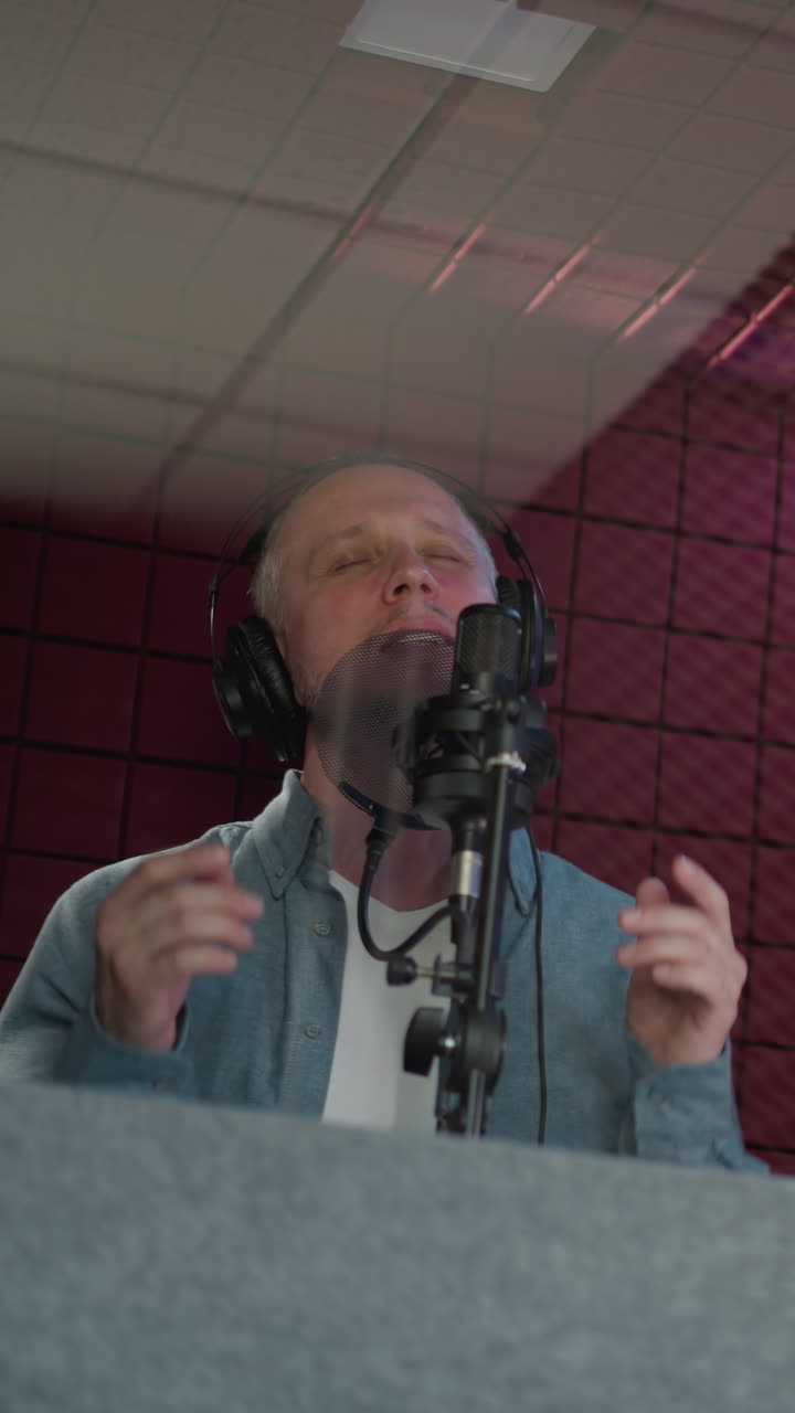 A man passionately sings in a recording studio, wearing black headphones and a blue long sleeve shirt. He performs with his mouth wide open, expressing emotion and intensity