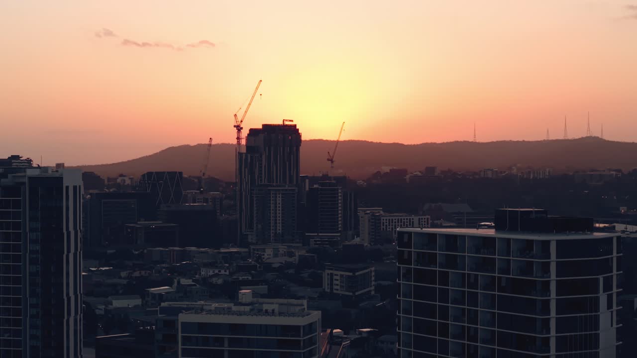 Drone sobe ao longo de edifícios modernos e altos, com silhuetas contra a luz quente do pôr do sol, revelando o desenvolvimento urbano, CBD Teneriffe Brisbane Queensland Austrália