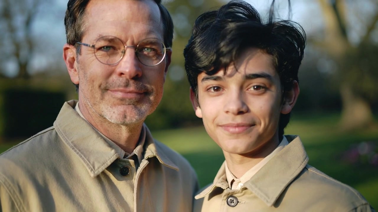 La telecamera si avvicina, padre e figlio in posa per un ritratto nel parco, che indossano cappotti beige e occhiali.
