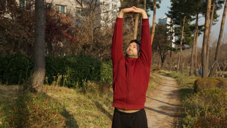 Hombre-Adulto-Medio-Haciendo-Ejercicio-Activo-Al-Aire-Libre-En-Un-Sendero-Del-Parque-Estirando-El-Torso-Bloqueando-El-Brazo-Por-Encima-De-La-Cabeza-Y-Saltando-Con-Calentamiento-De-Hombros-Rotaciones-Al-Aire-Libre-En-El-Parque-De-Apartamentos-En-Otoño---Cámara-Lenta