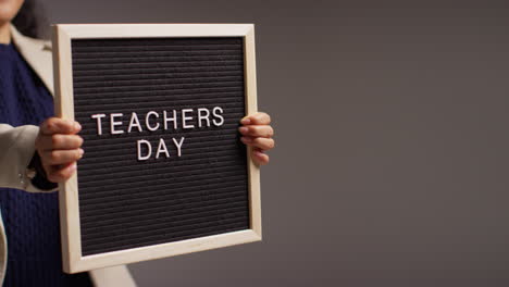 Close-Up-Studio-Shot-Of-Female-Teacher-Standing-Against-Grey-Background-Holding-Notice-Board-Reading-Teachers-Day-2