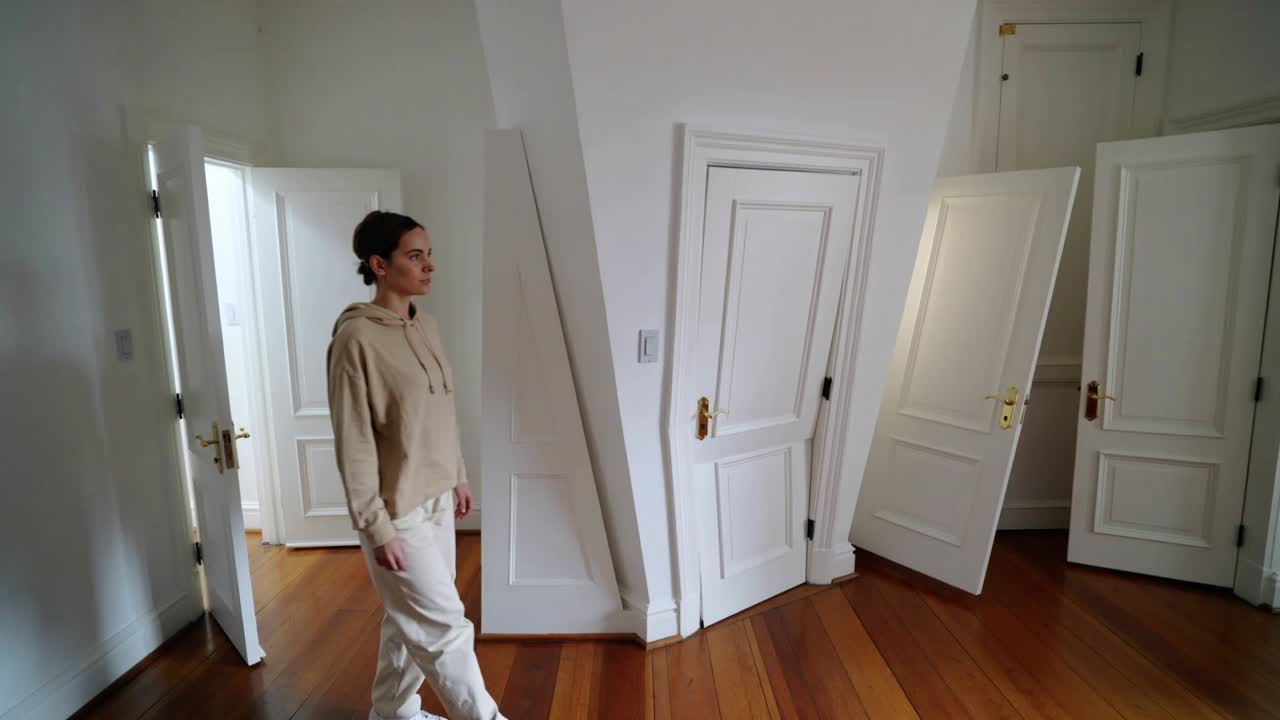 Donna in felpa con cappuccio beige che entra da sinistra, attraversando la stanza su pavimento in legno, oltrepassando porte bianche.