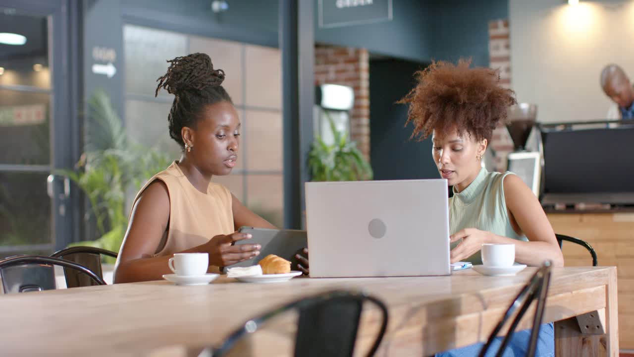 African American women opening laptop and gesturing, sharing tablet while planning project at cafe