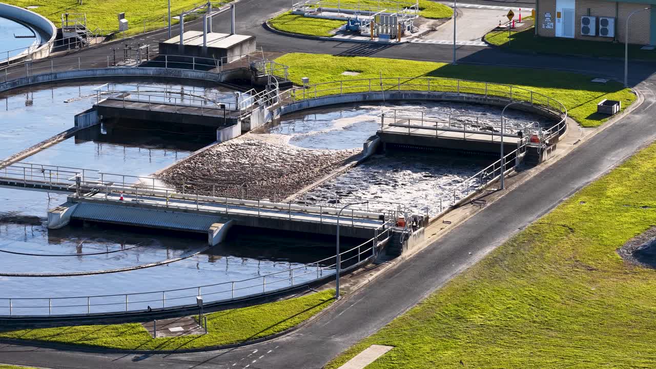 Aerial footage pans across a large wastewater treatment plant with settling tanks, green lawns, and industrial structures under bright natural daylight