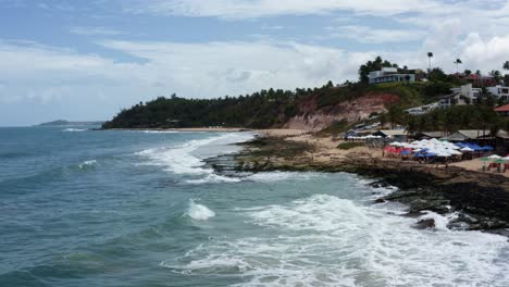 Toma-Aérea-De-Drones-De-La-Península-Tropical-De-Tibau-Do-Sul-Con-Olas-Rompiendo-En-Rocas-Cubiertas-De-Algas,-Turistas-Disfrutando-De-La-Sombra-Bajo-Coloridos-Paraguas-Y-Grandes-Acantilados-En-Río-Grande-Do-Norte,-Brasil