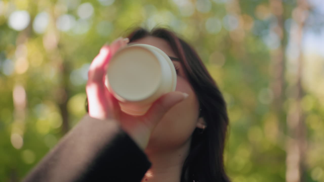 Medium shot of autumn visitor strolling through arboretum, sipping morning tea, eyes lifting toward light with quiet contentment, soft sun beam across face, blur forest bokeh behind