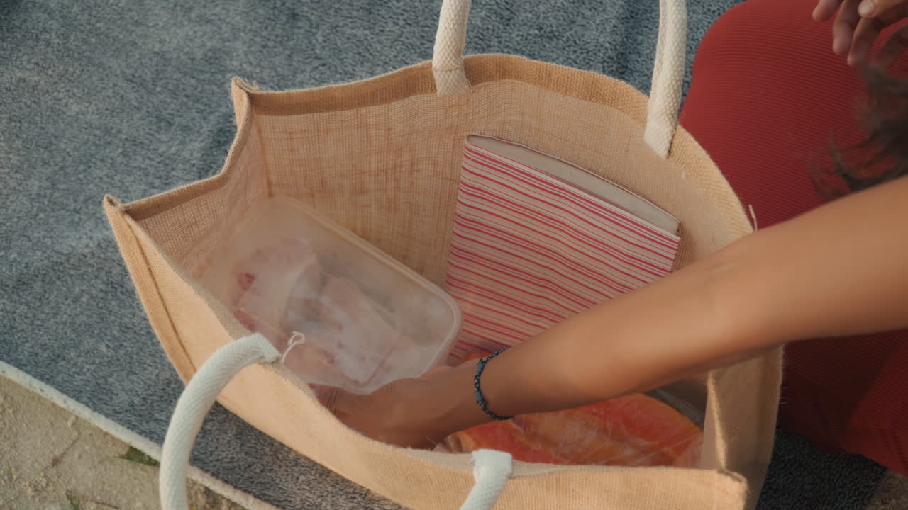 Borsa di paglia piena di essenziali per il picnic, donna che mette la mano nella borsa per prendere un contenitore sigillato e degli snack, primo piano di mani che organizzano gli scomparti, manica di un vestito rosso visibile, preparazione informale per la spiaggia.