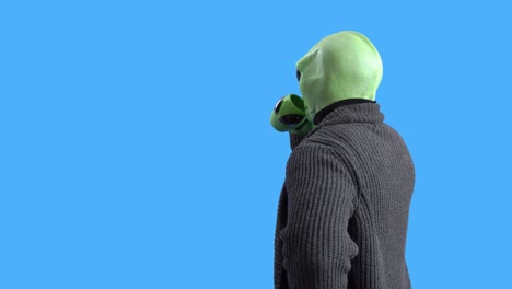 man in alien costume, warm knitted jacket, with funny themed mug in his hand pretends to wipe the window with sleeve and drink hot beverage against a blue chroma key. preparation for halloween party