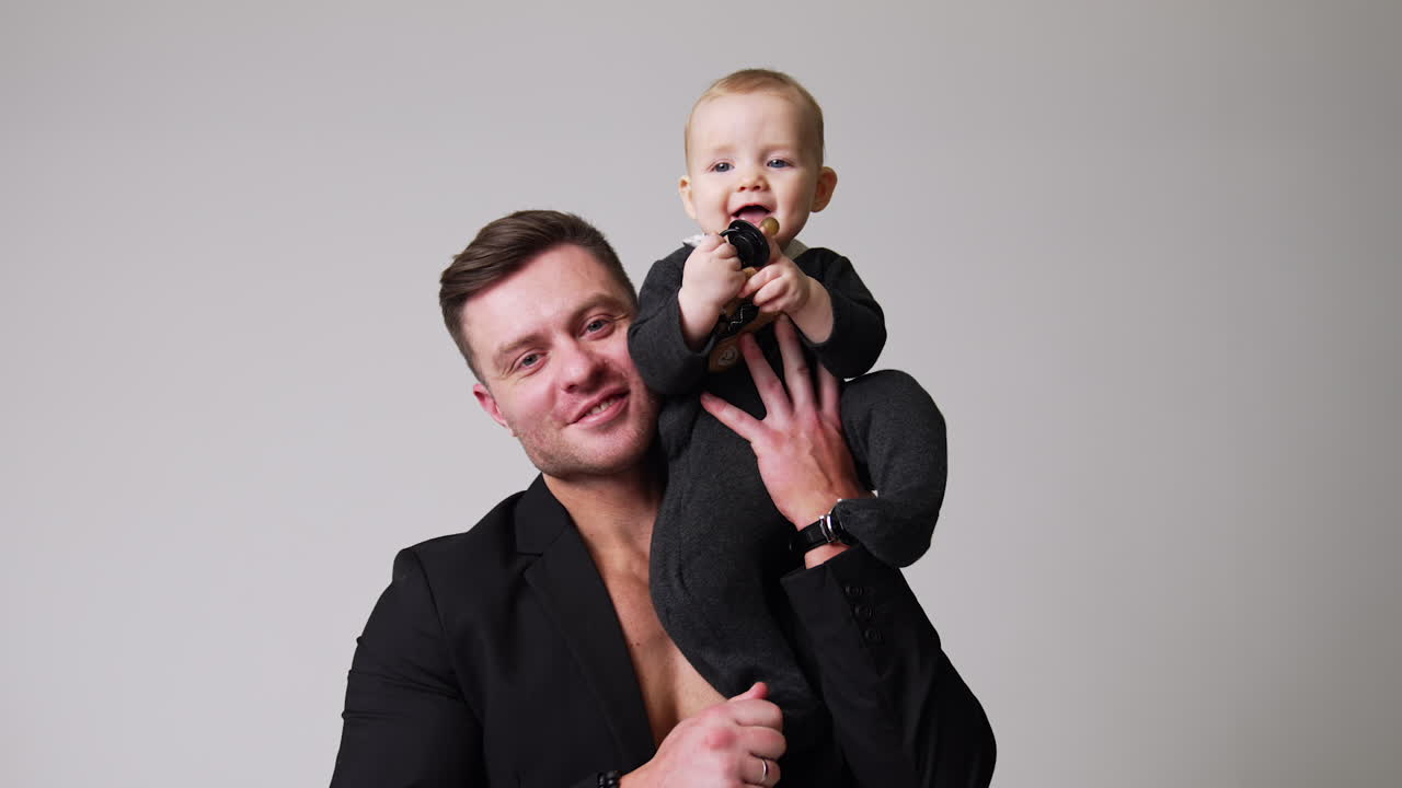 Loving father holding a little baby on his shoulder. Happy little child pushes the pacifier to his mouth. Studio footage. White backdrop.