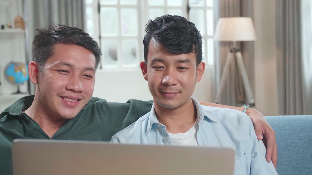 Portrait Of Asian Gentle Gay Couple Using Laptop Computer, While Sitting On A Couch In Cozy Stylish Apartment. Males Looking Each Other