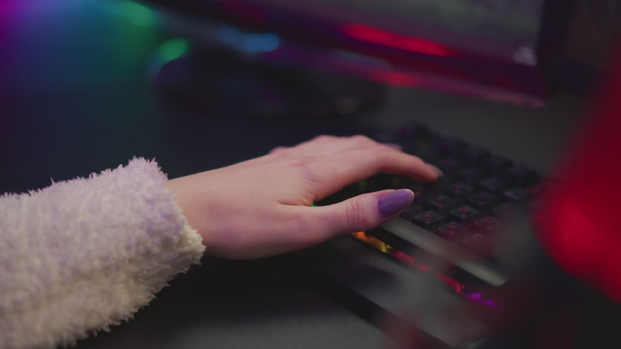 Hand view of gamer with polished nails actively typing on glowing mechanical keyboard while playing game, partial view of gaming monitor visible in background