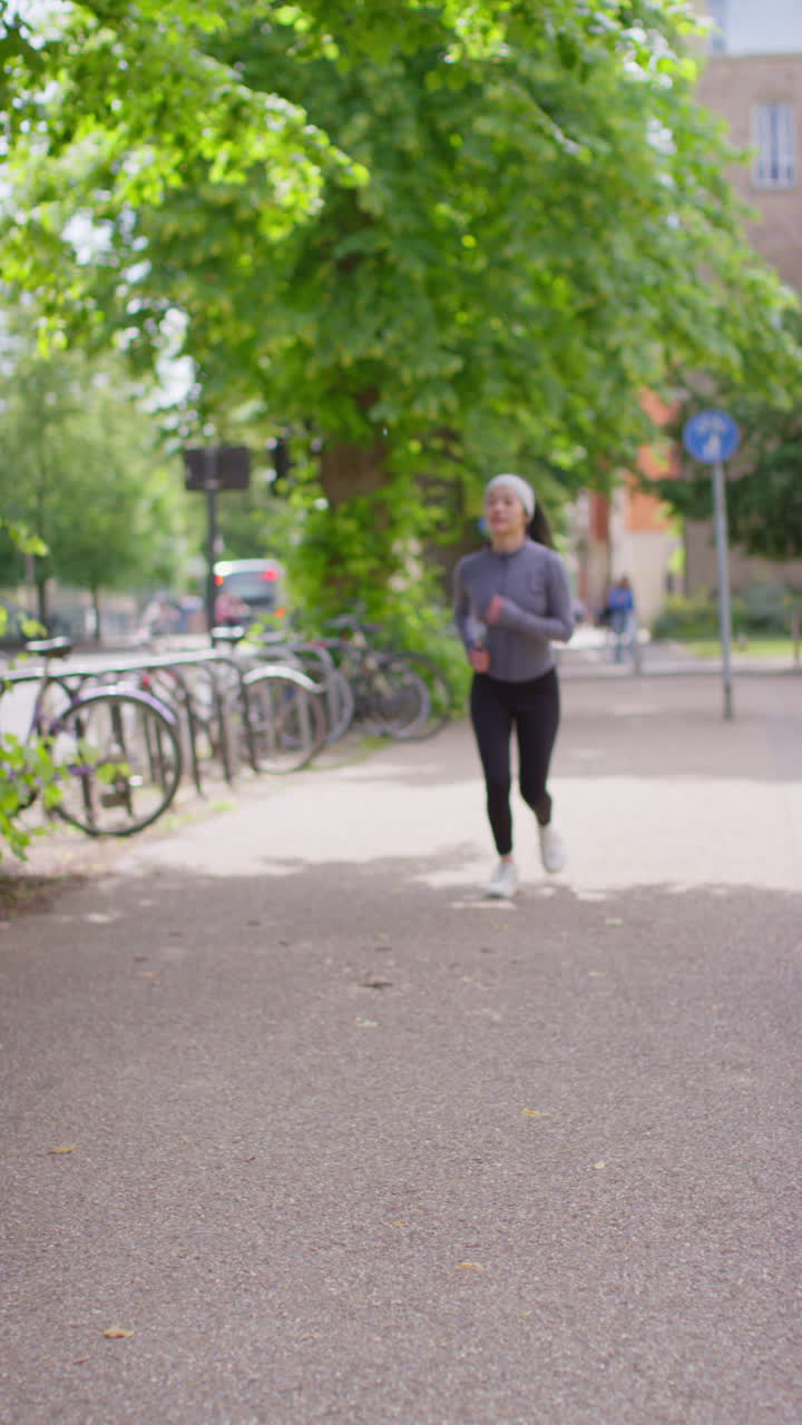 Vertical Video Of Young Woman Exercising Running Along Path Through City Park Wearing Wireless Earbuds