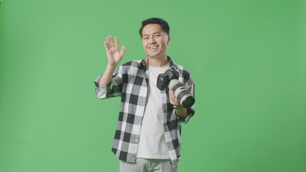 아시아 사진작가 가 스튜디오 의 녹색 스크린 배경 에 서 있는 동안 카메라 에 있는 사진 을 보고 손 을 흔들고 있다