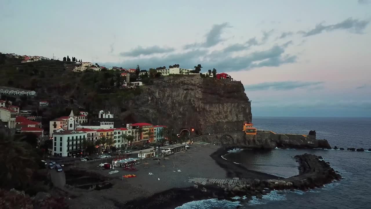 Basking in the warm glow of dusk, Ponta do Sol in Madeira, Portugal, features colorful buildings perched on cliffs overlooking the tranquil Atlantic Ocean, flying forward establishing drone shot
